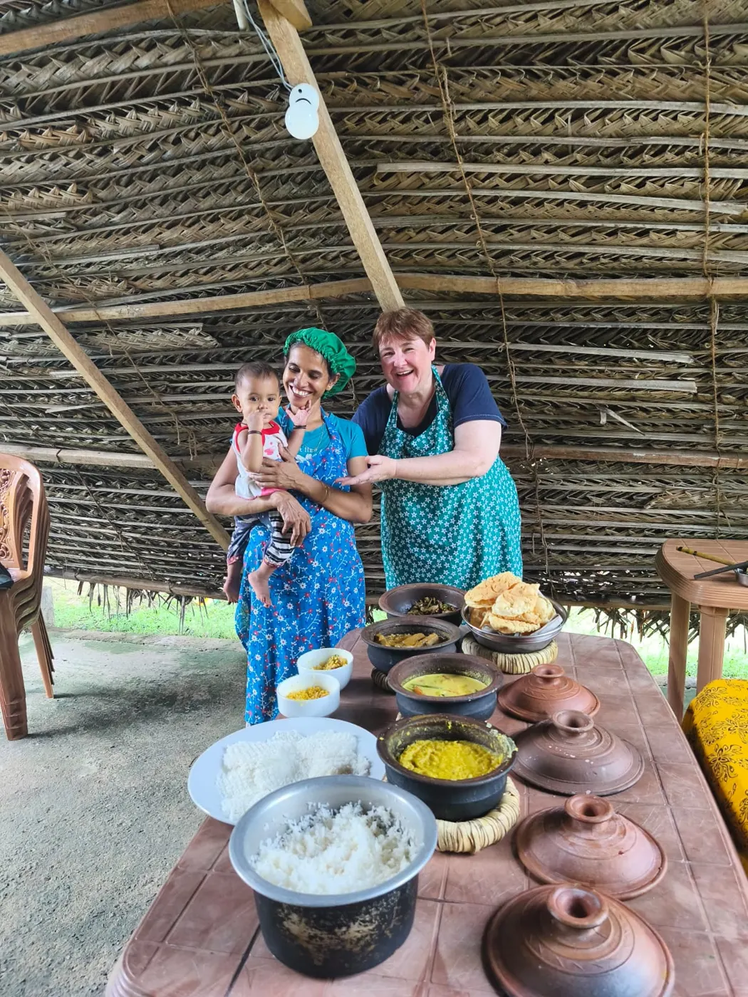 Nimali with her baby presenting a full traditional Sri Lankan rice and curry spread in her palm-leaf kitchen