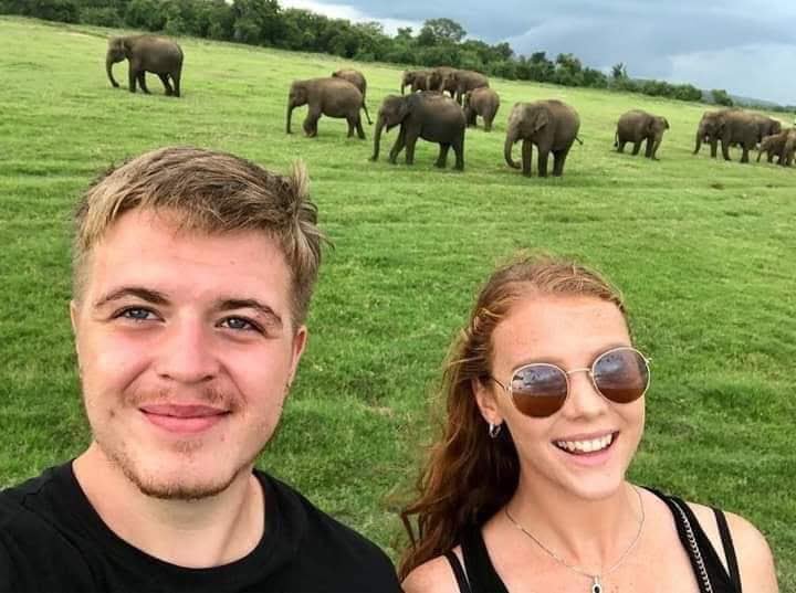 Two safari guests smiling with a herd of wild elephants grazing behind them at Kaudulla